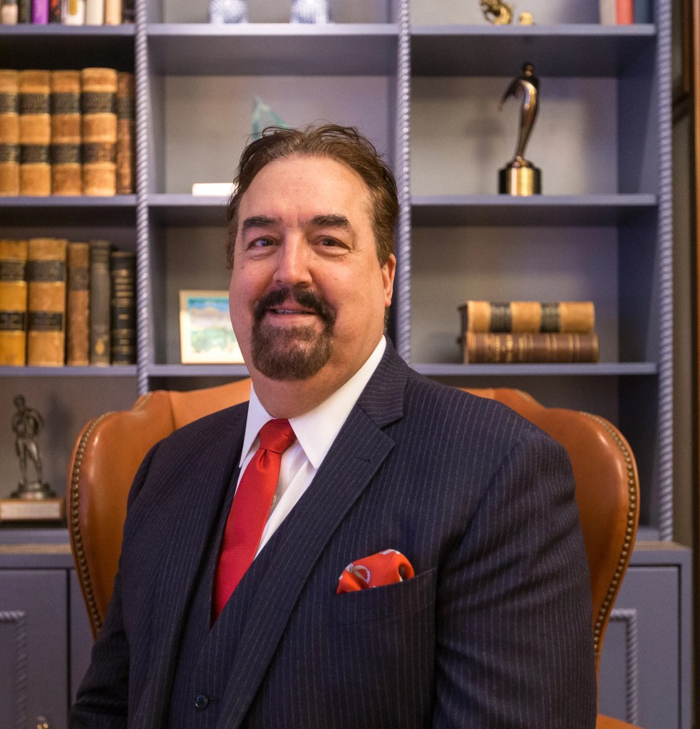 A well-dressed middle-aged or older man sits in an office, characterized by a dark pinstripe suit paired with a crisp whit...
